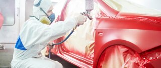 Ein Mann in Schutzkleidung sprüht roten Lack auf ein Auto in einer Werkstatt. Das Fahrzeug ist teilweise mit Folie abgedeckt.