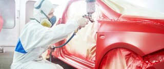 Ein Mann in Schutzkleidung sprüht roten Lack auf ein Auto in einer Werkstatt. Das Fahrzeug ist teilweise mit Folie abgedeckt.