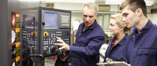 Ein erfahrener Fachmann erklärt jungen Mitarbeitern die Bedienung einer Maschinensteuerung in einer Werkstatt.