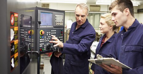 Ein erfahrener Fachmann erklärt jungen Mitarbeitern die Bedienung einer Maschinensteuerung in einer Werkstatt.