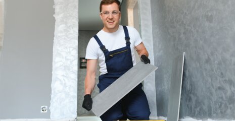 Ein Handwerker in blauer Latzhose und T-Shirt lächelt, während er eine Fliese in der Hand hält. Der Raum ist frisch renoviert.