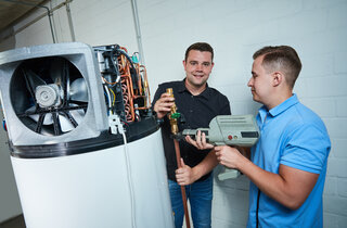 Zwei Männer arbeiten an einer technischen Anlage. Einer hält ein Glas, während der andere ein Werkzeug benutzt. Sie scheinen in einer Werkstatt zu sein.