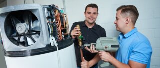 Zwei Männer arbeiten an einer technischen Anlage. Einer hält ein Glas, während der andere ein Werkzeug benutzt. Sie scheinen in einer Werkstatt zu sein.