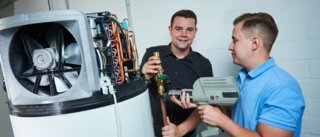 Zwei Männer arbeiten an einer technischen Anlage. Einer hält ein Glas, während der andere ein Werkzeug benutzt. Sie scheinen in einer Werkstatt zu sein.