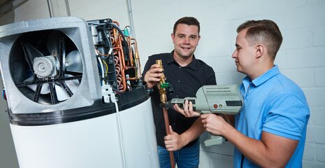 Zwei Männer arbeiten an einer technischen Anlage. Einer hält ein Glas, während der andere ein Werkzeug benutzt. Sie scheinen in einer Werkstatt zu sein.