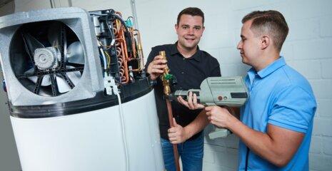 Zwei Männer arbeiten an einer technischen Anlage. Einer hält ein Glas, während der andere ein Werkzeug benutzt. Sie scheinen in einer Werkstatt zu sein.