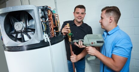 Zwei Männer arbeiten an einer technischen Anlage. Einer hält ein Glas, während der andere ein Werkzeug benutzt. Sie scheinen in einer Werkstatt zu sein.