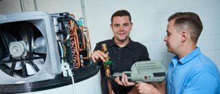 Zwei Männer arbeiten an einer Heizungsanlage. Einer hält ein Werkzeug, der andere zeigt auf ein Ventil. Die Atmosphäre wirkt industriell und technisch.
