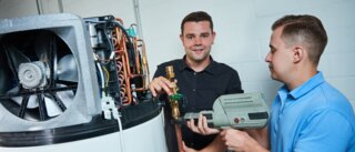 Zwei Männer arbeiten an einer Heizungsanlage. Einer hält ein Werkzeug, der andere zeigt auf ein Ventil. Die Atmosphäre wirkt industriell und technisch.