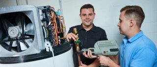 Zwei Männer arbeiten an einer Heizungsanlage. Einer hält ein Werkzeug, der andere zeigt auf ein Ventil. Die Atmosphäre wirkt industriell und technisch.