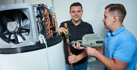 Zwei Männer arbeiten an einer Heizungsanlage. Einer hält ein Werkzeug, der andere zeigt auf ein Ventil. Die Atmosphäre wirkt industriell und technisch.