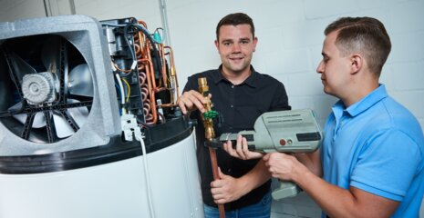 Zwei Männer arbeiten an einer Heizungsanlage. Einer hält ein Werkzeug, der andere zeigt auf ein Ventil. Die Atmosphäre wirkt industriell und technisch.