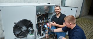 Zwei Techniker arbeiten an einer Klimaanlage. Einer erklärt und zeigt Messgeräte, während der andere an den Leitungen arbeitet.