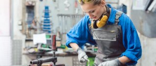 Eine junge Frau in Arbeitskleidung steht an einer Werkbank, konzentriert bei der Arbeit mit Werkzeugen. Sicherheitsbrille und Ohrenschutz tragen sie zum Schutz.