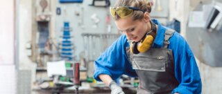 Eine junge Frau in Arbeitskleidung steht an einer Werkbank, konzentriert bei der Arbeit mit Werkzeugen. Sicherheitsbrille und Ohrenschutz tragen sie zum Schutz.