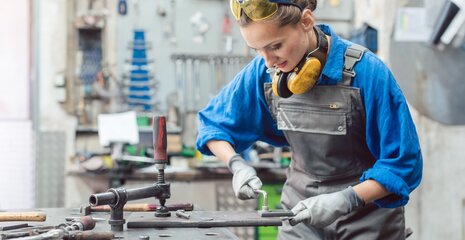 Eine junge Frau in Arbeitskleidung steht an einer Werkbank, konzentriert bei der Arbeit mit Werkzeugen. Sicherheitsbrille und Ohrenschutz tragen sie zum Schutz.