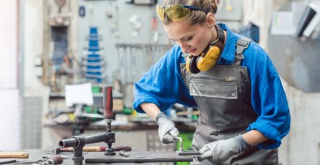 Eine junge Frau in Arbeitskleidung steht an einer Werkbank, konzentriert bei der Arbeit mit Werkzeugen. Sicherheitsbrille und Ohrenschutz tragen sie zum Schutz.