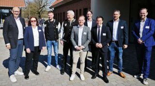 Die Gruppe von Personen steht vor einem Gebäude an einem sonnigen Tag. Sie lächeln und sind in verschiedenen, formellen Outfits gekleidet.