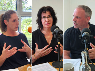 Das Bild zeigt drei Personen, die in einem Gespräch sind. Sie scheinen engagiert zu diskutieren, umgeben von Mikrofonen und Notizen.