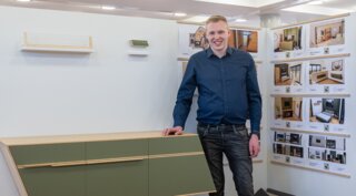 Der junge Mann steht neben einer modernen, grünen Wandkonsole. Im Hintergrund sind Designpläne und Bilder zu sehen.