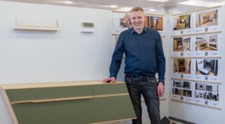 Der junge Mann steht neben einer modernen, grünen Wandkonsole. Im Hintergrund sind Designpläne und Bilder zu sehen.