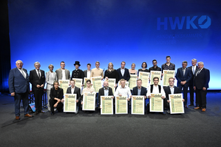 Auf dem Bild stehen mehrere Personen auf einer Bühne, sie halten Urkunden in den Händen und lächeln. Im Hintergrund ist die Bezeichnung "HWK" zu sehen.
