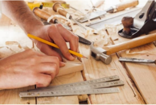 Eine Hand hält einen Bleistift, während auf einem Holzstück gemessen wird. Werkzeuge und Holzspäne umgeben die Arbeitsfläche.
