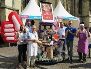 Auf dem Bild stehen mehrere Personen vor einem Stand. Sie lächeln und halten Brot. Im Hintergrund sind Zelte und eine große Daumen-hoch-Dekoration zu sehen.