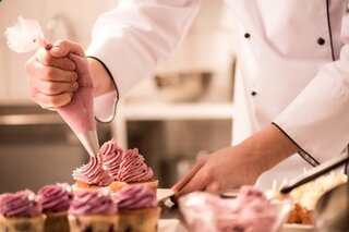 Ein Konditor verziert Cupcakes mit rosa Zuckerguss. Die Kreativität und Präzision sind deutlich sichtbar. Ein köstlicher Anblick!