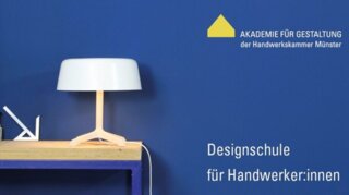 Das Bild zeigt eine moderne Tischlampe auf einem Holztisch vor einer blauen Wand. Daneben stehen Werkzeuge und Materialien für Handwerker:innen.