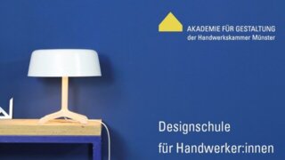 Das Bild zeigt eine moderne Tischlampe auf einem Holztisch vor einer blauen Wand. Daneben stehen Werkzeuge und Materialien für Handwerker:innen.