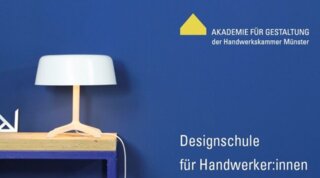 Das Bild zeigt eine moderne Tischlampe auf einem Holztisch vor einer blauen Wand. Daneben stehen Werkzeuge und Materialien für Handwerker:innen.